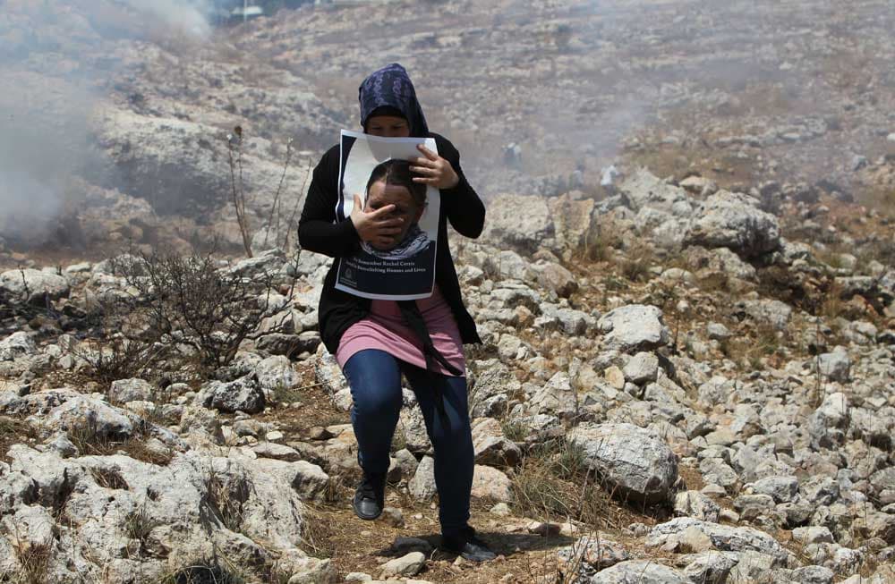 سيدة تهرب من القوات الاسرائيلية وهي تحمل صورة راشيل كوري الناشطة الأمريكية التي دهست من قبل جرافة  خلال تظاهرة في غزة في عام 200