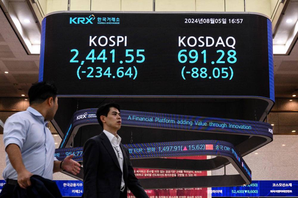 People walk under a monitor displaying the benchmark Kospi index and the Korean Securities Dealers Automated Quotations (KOSDAQ) after the close of trading in Seoul on August 5, 2024. Seoul's stocks plunged more than eight percent at closing, after weak US jobs data fanned fears of a recession in the world's top economy and boosted bets on several Federal Reserve interest rate cuts. (Photo by ANTHONY WALLACE / AFP)