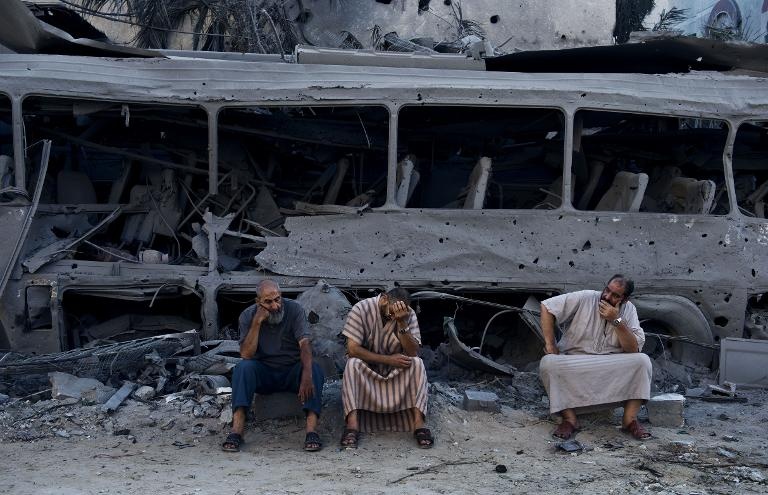 ثلاثة فلسطينيين يجلسون أمام حافلة ومنازل مدمرة مقابل منزل سوي بالارض بعد استهدافه بغارات إسرائيلية في غزة